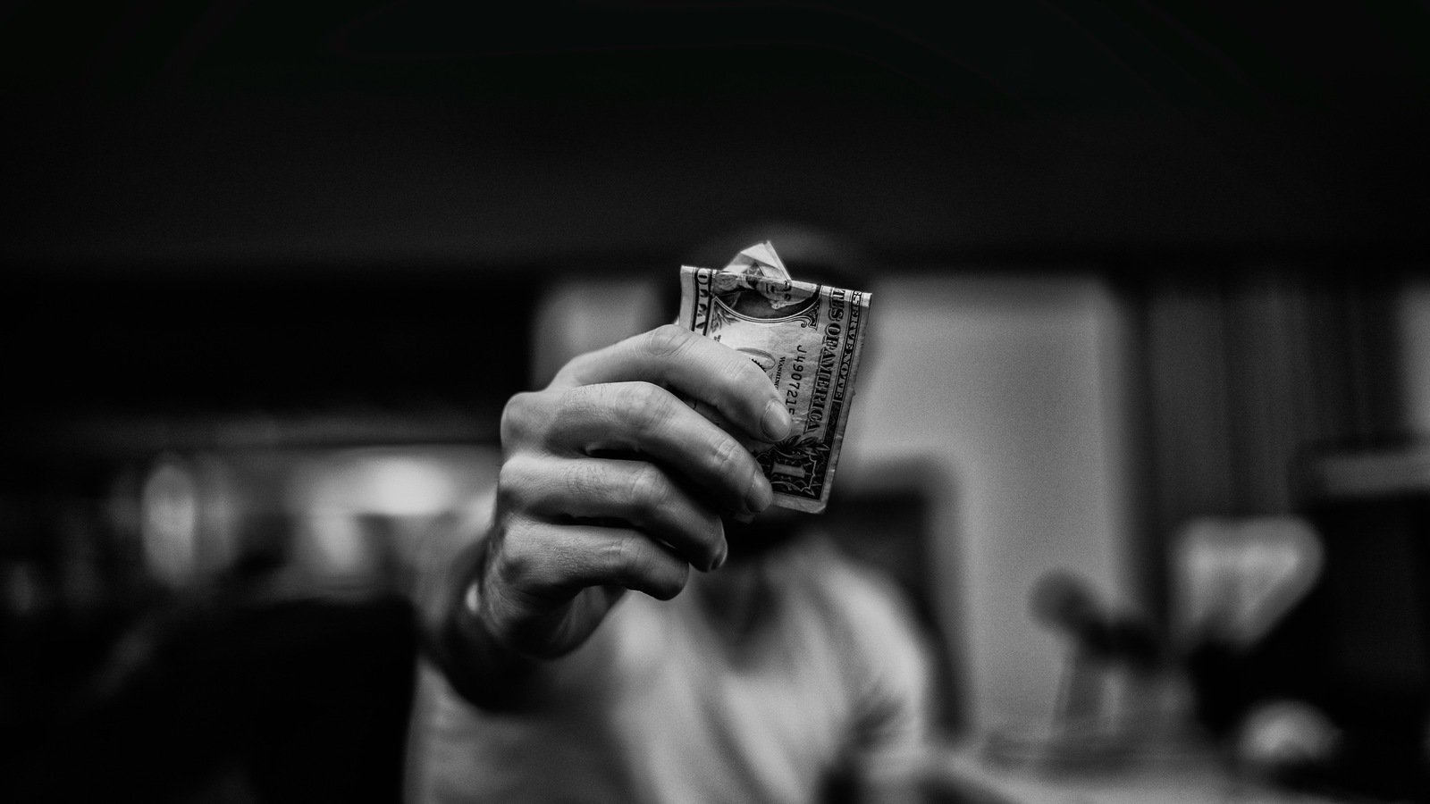 A blurred black and white image of a person holding a crumpled one-dollar bill forward. How much should you spend on Google Ads- Learn budgeting strategies from NewFrame Digital